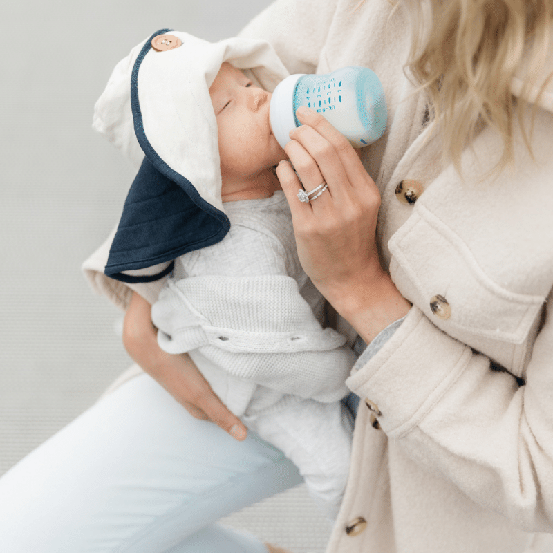 A woman bottle feeds her baby on a bench, using The Mobobaby Breastfeeding Hat, An innovative new, breathable nursing cover, it is a great hooter hider so you can enjoy covered goods. A unique and best baby gift, it's a perfect breastfeeding cover up. 